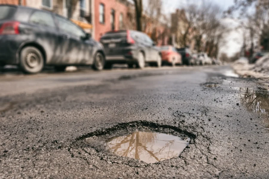 A large water-filled pothole in a cracked asphalt street with cars parked along the curb in the background.